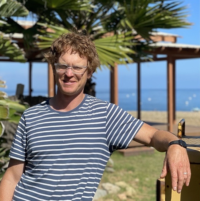 A person wearing glasses and a striped shirt stands outdoors by a palm tree and a building near the beach, evoking the laid-back vibe of summer camp on Catalina Island.